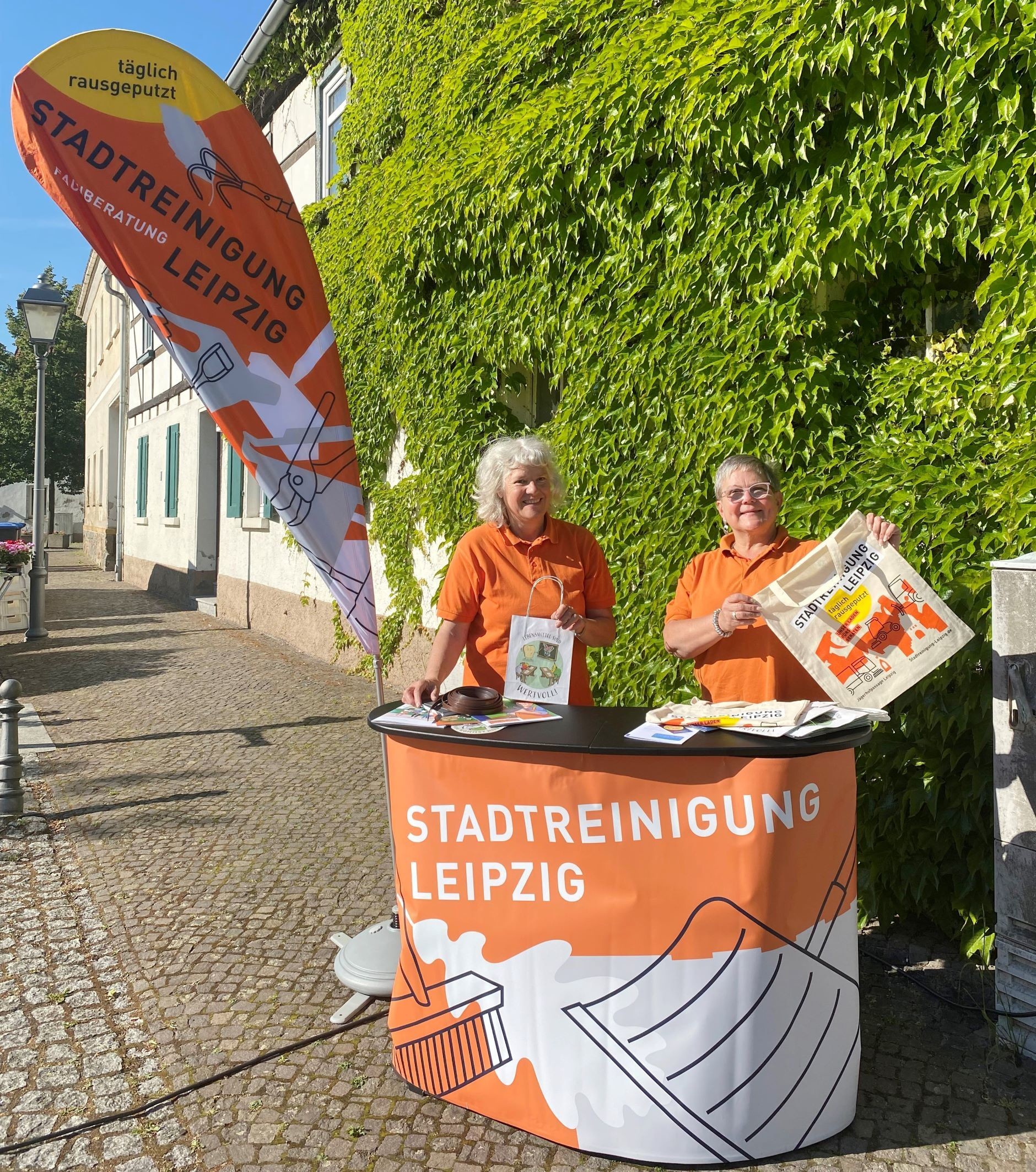 Zwei Frauen mit orangem T-Shirt stehen hinter einem Tresen, auf dem "Stadtreinigung Leipzig" steht.