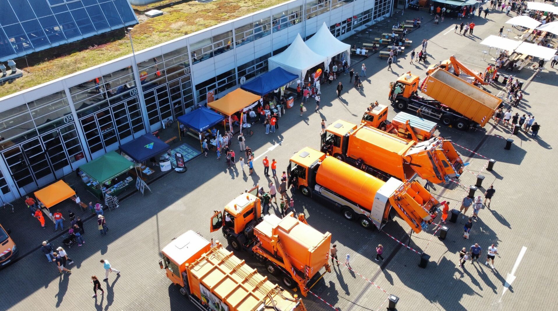 Aus Drohnenperspektive ist das Betriebsgel&auml;nde der Stadtreinigung Leipzig zusehen. Orange Fahrzeuge der Stadtreinigung sind aufgereiht. Gegen&uuml;ber stehen bunte Zelte und dazwischen laufen einige Menschen.