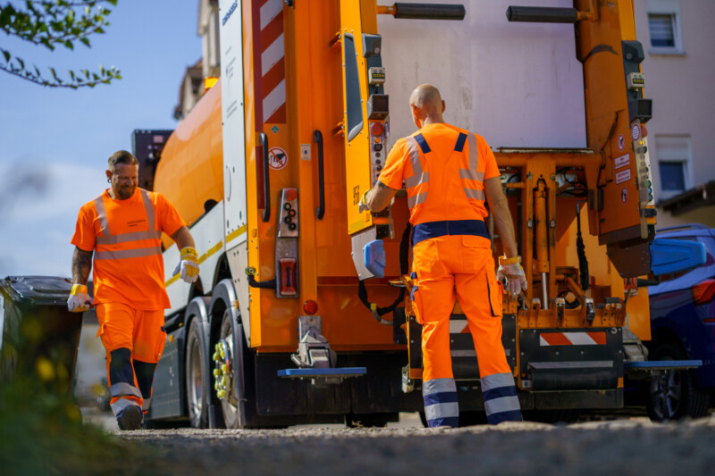 Ein Müllwerker holt eine schwarze Restabfalltonne und ein zweiter Müllwerker steht am Abfallsammelfahrzeug, um sie anzunehmen