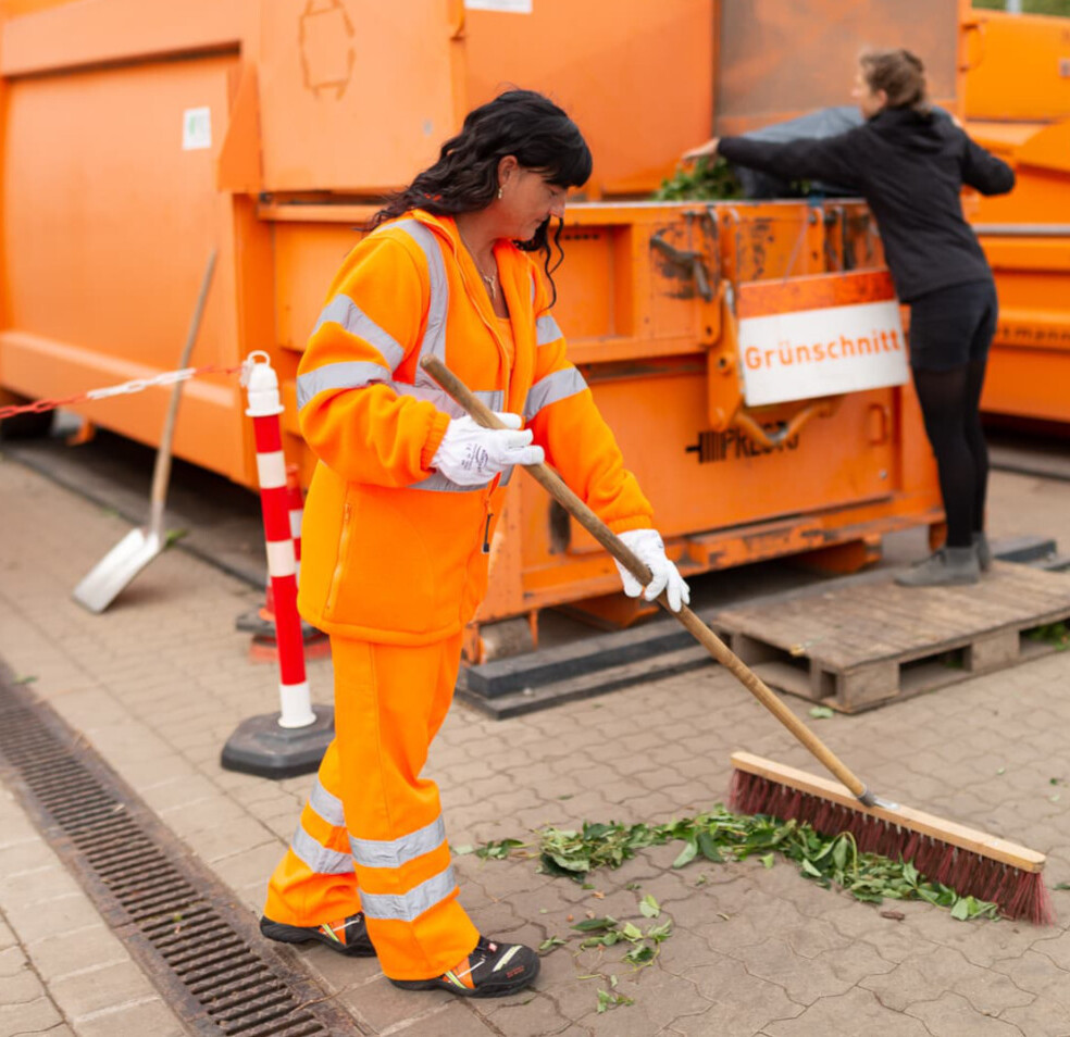 Eine Frau gibt Gr&uuml;nschnitt in den Container. Eine Mitarbeiterin fegt lose Bl&auml;tter davor zusammen.