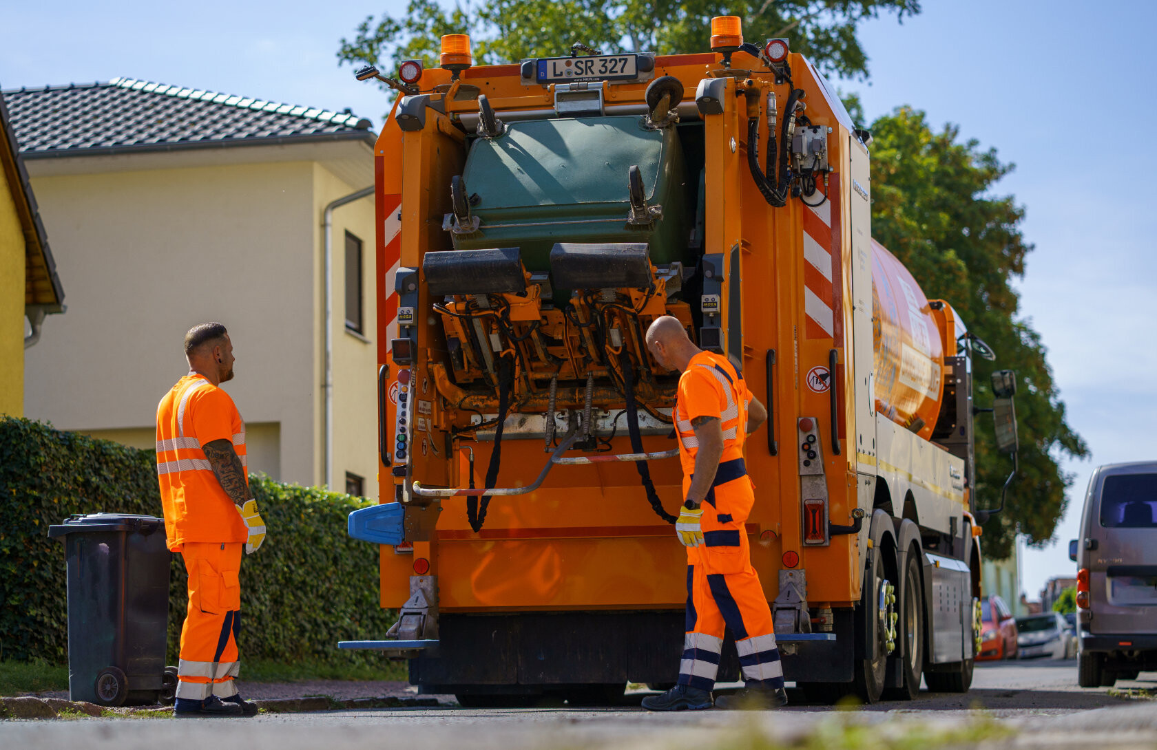 Zwei M&uuml;llwerker kippen eine gro&szlig;e Restabfalltonne in ein oranges Sammelfahrzeug ab. Sie stehen daf&uuml;r hinter dem Fahrzeug, das man von hinten sieht.