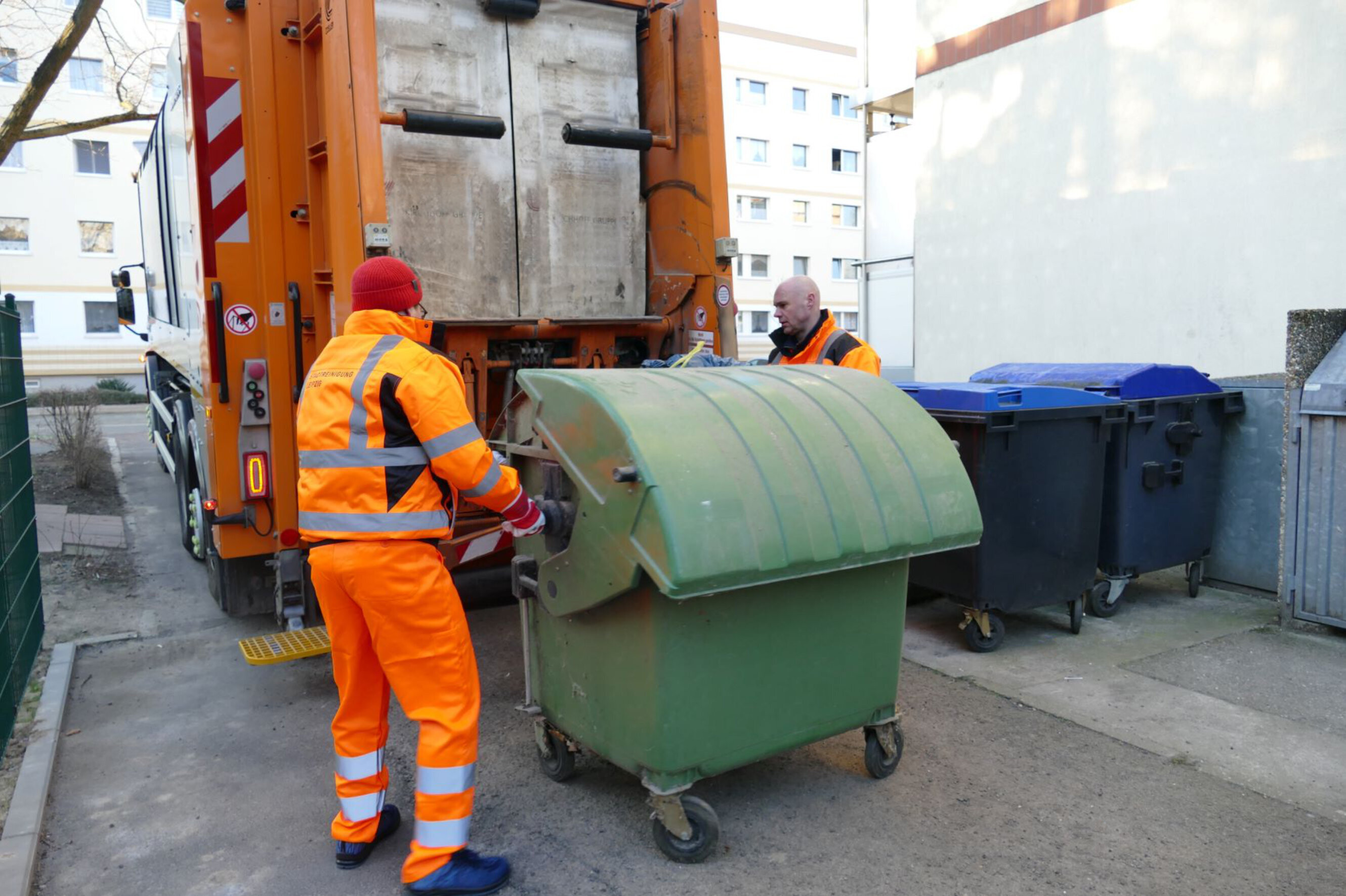Zwei M&uuml;llwerker stehen mit einer Abfalltonne hinter einem Abfallsammelfahrzeug. Die Tonne wird langsam vom Fahrzeug zum Abkippen angehoben