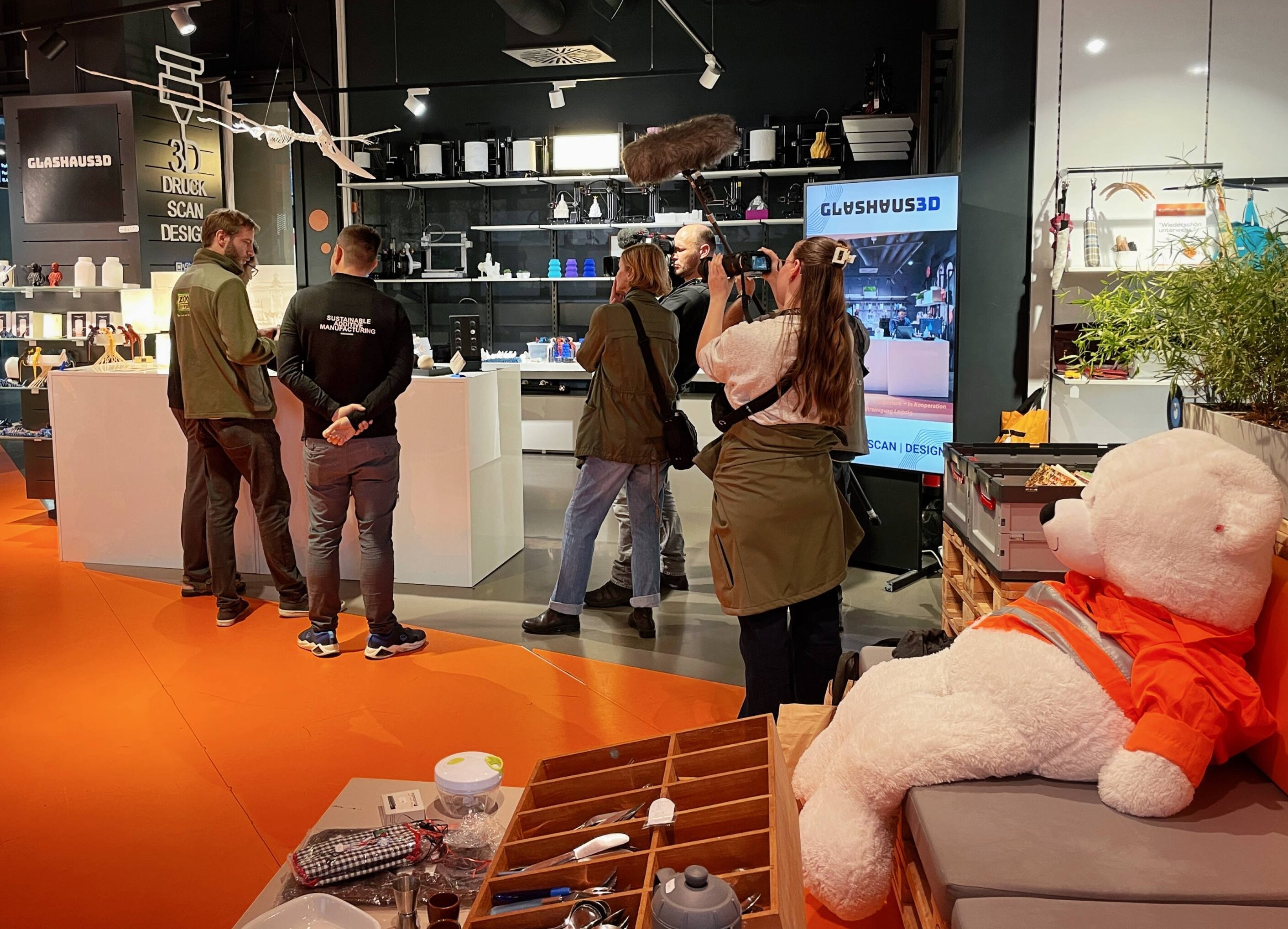 Ein Kameramann, ein Tontechniker, eine Reporterin, zwei M&auml;nner von der Firma Glashaus 3D, ein Tierpfleger aus dem Leipziger Zoo und eine Praktikantin aus dem Leipziger Zoo stehen im Wiedersch&ouml;n am Stand der Firma Glaushaus 3D und filmen die neuen Futterger&auml;te. Im Vordergrund am rechten Rand des Bildes sitzt ein gro&szlig;er Teddy aus dem Wiedersch&ouml;n.