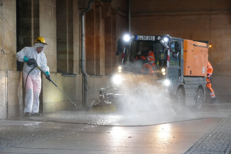 Person links mit Nassreiniger spritzt den Boden mit Wasser ab. Kleinkehrmaschine, rechts, fährt über die gereinigte Stelle