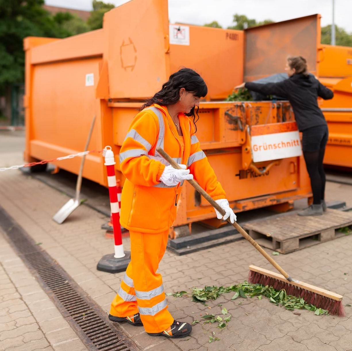 Eine Mitarbeiterin fegt vorm Grünschnittcontainer Laub zusammen.