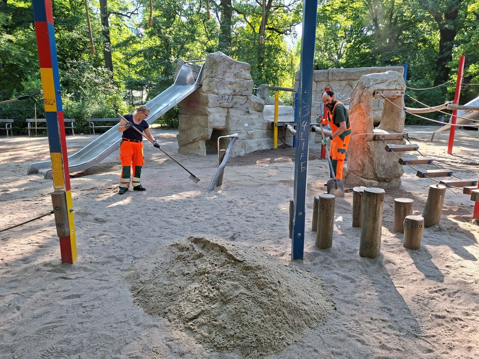2 Mitarbeitende f&uuml;llen Sand auf einem Spielplatz auf.
