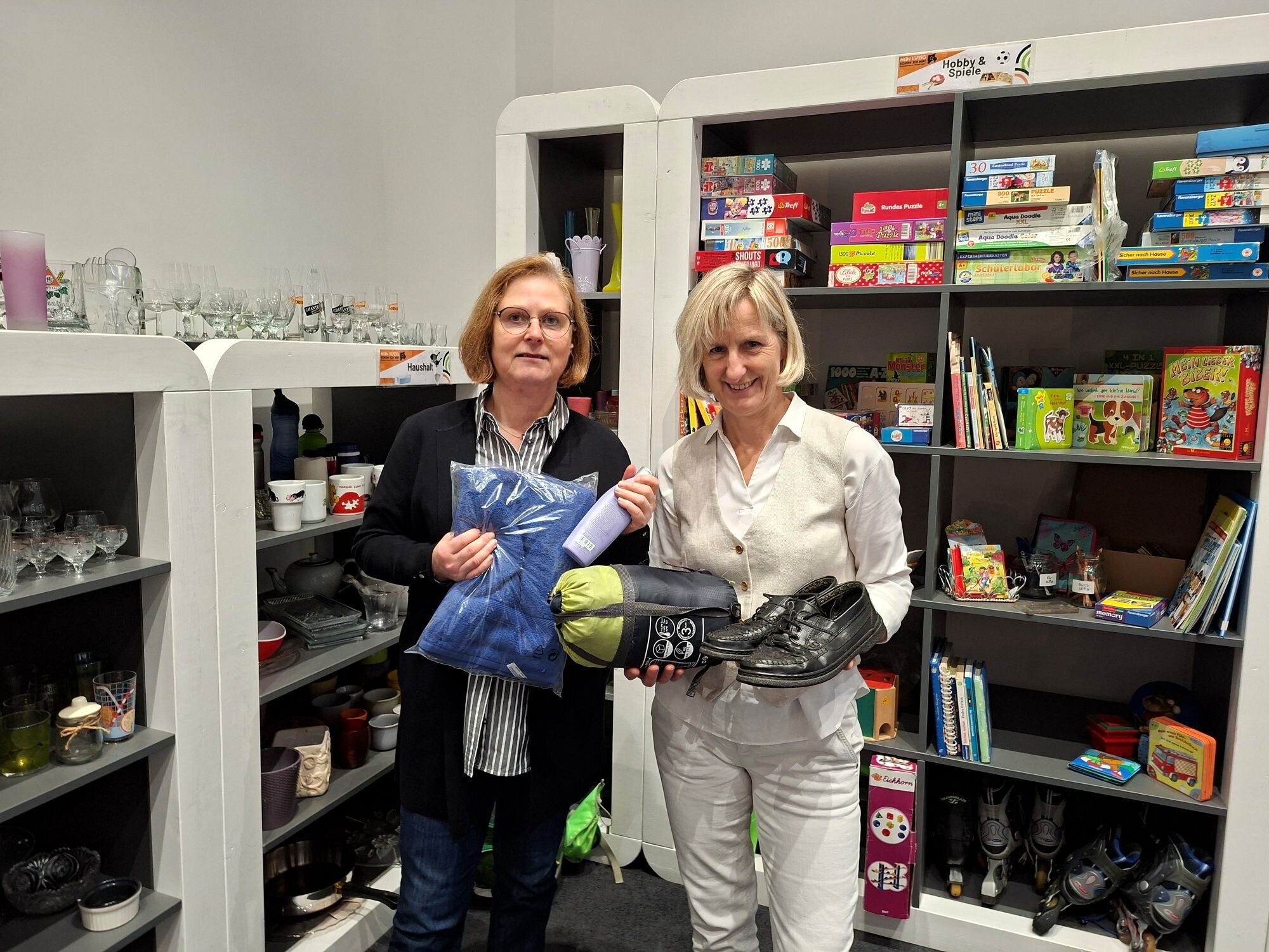 Martina Kador Probst, Leiterin des Sozialamts Leipzig, steht links und hält ein Handtuch und ein Shampoo in der Hand. Kaufmännische Betriebsleiterin der Stadtreinigung Leipzig, steht rechts von ihr und hält Schuhe und einen Schlafsack.. Elke Bröcker,