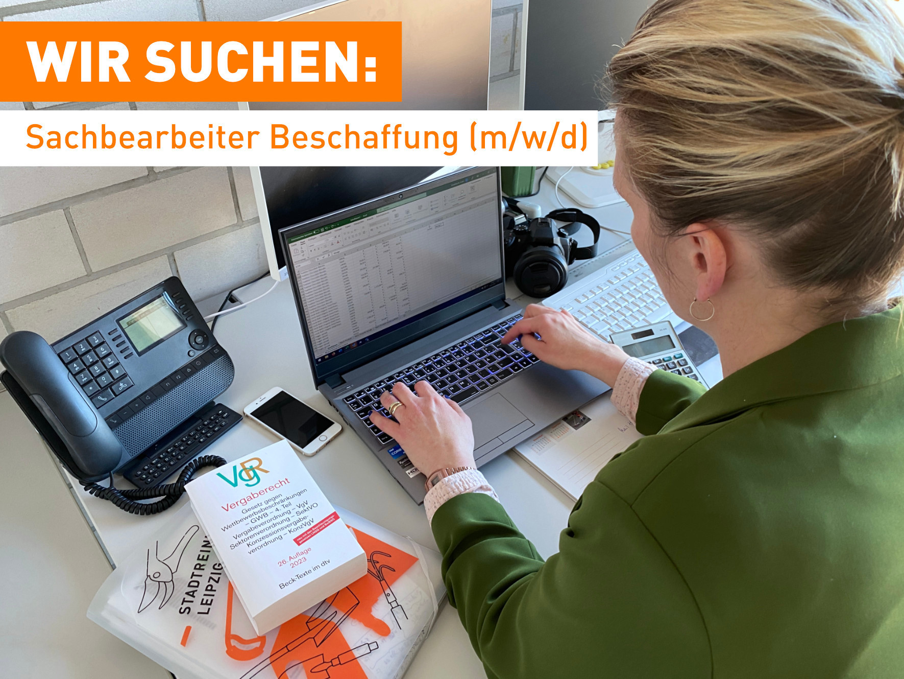 Eine blonde Frau Arbeitet am Laptop. neben ihr liegen ein Regelwerk, Handy, Telefon, Kamera, Taschenrechner.