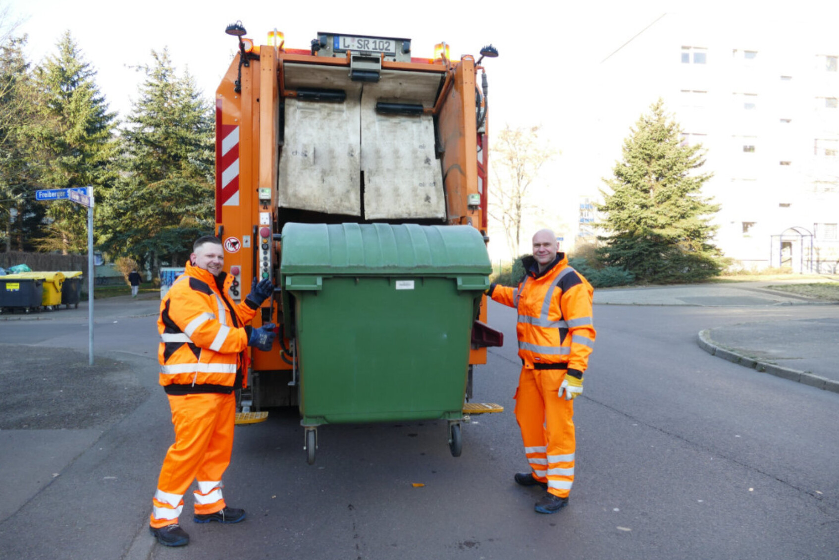 Zwei Müllarbeiter stehen mit einer Abfalltonne hinter einem Abfallsammelfahrzeug. Die Tonne wird langsam vom FAhrzeug zum Abkippen angehoben