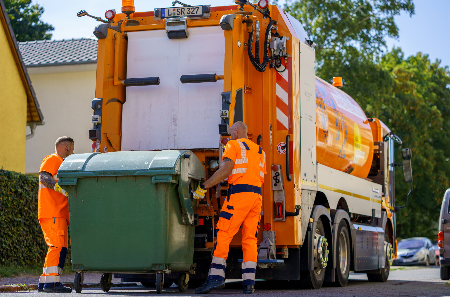Zwei M&uuml;llwerker bereiten gemeinsam eine gro&szlig;e gr&uuml;ne Restabfalltonne zum Auskippen in das Abfallsammelfahrzeug vor. Daf&uuml;r stehen sie hinter dem Fahrzeug.