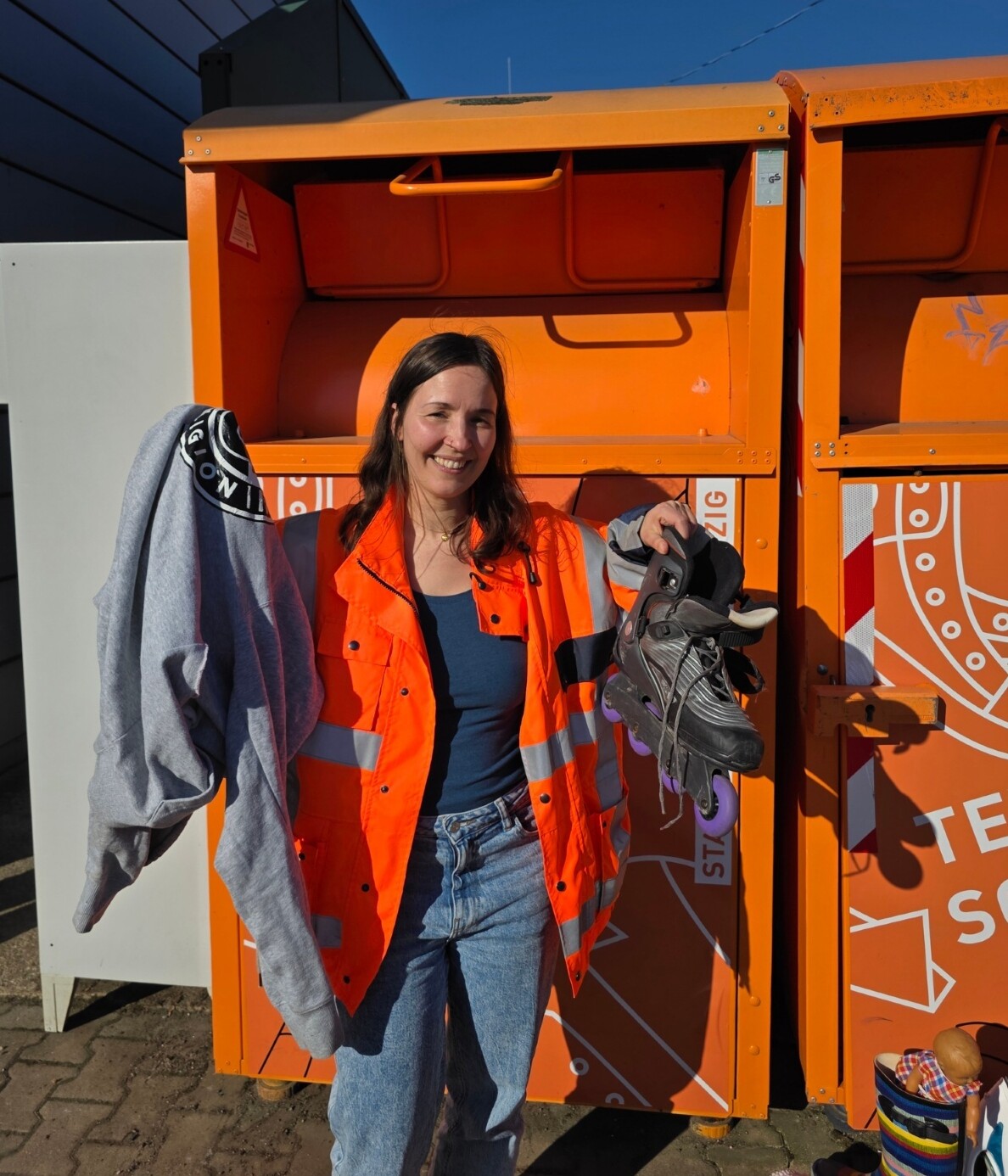 Eine Frau in Warnjacke steht vor den orangen Altkleidercontainer und h&auml;lt in ihrer rechten Hand einen Pullover und in ihrer linken Hand Inline-Skater hoch.