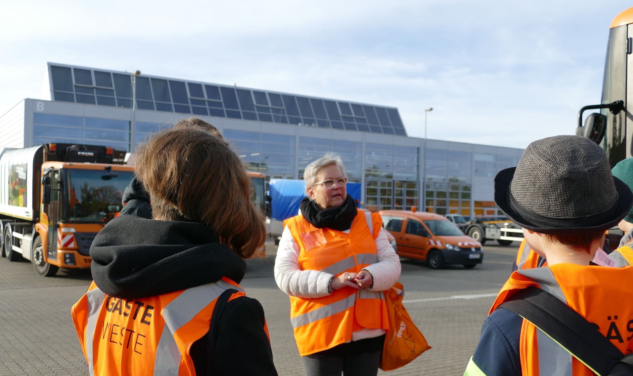 Personen stehen in orangenen Warnwesten auf dem Betriebsgel&auml;nde der Stadtreinigung. Eine Mitarbeiterin der Stadtreinigung erz&auml;hlt etwas zum Betriebsobjekt. Im Hintergrund sind Abfallsammelfahrzeuge zu sehen.